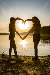 Full length of friends making heart shape with hands while standing at lakeshore during sunset - OJF00394