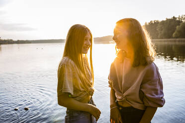 Smiling friends standing together against lake during sunset - OJF00397