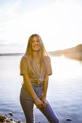 Portrait of happy girl standing at lakeshore against sky during sunset - OJF00398