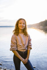 Smiling girl standing at lakeshore against sky during sunset - OJF00399