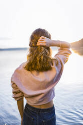 Rückansicht eines Mädchens mit Hand im Haar, das bei Sonnenuntergang auf einen See blickt - OJF00400