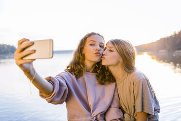 Freunde spitzen die Lippen, während sie ein Selfie mit dem Smartphone am See machen - OJF00401