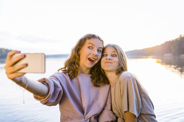 Happy friends making faces while taking selfie on smart phone against lake - OJF00403