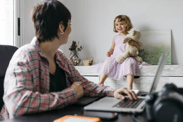 Mother working at home, daughter sitting with teddy bear on sideboard - JRFF04361