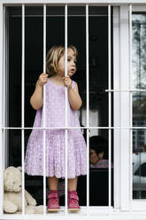 Girl standing at open window looking through window grate - JRFF04368