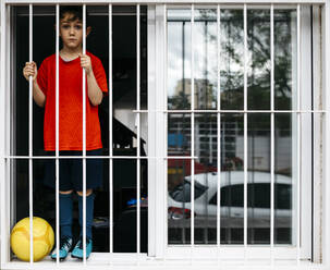 Boy with soccer ball at open window looking through window grate - JRFF04369