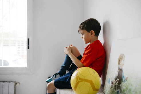 Sad boy with soccer ball sitting on sideboard - JRFF04375