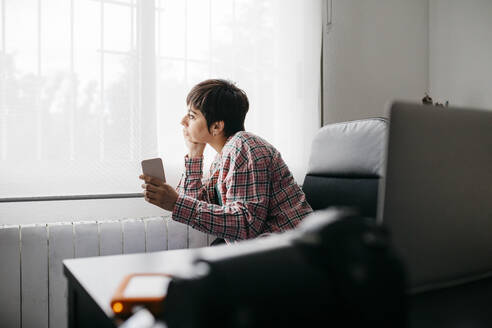 Female freelancer looking out of the window at home, holding smartphone - JRFF04379