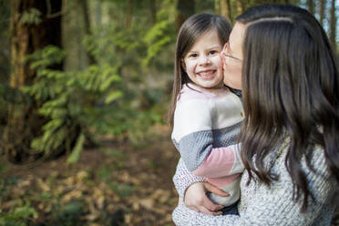 Mother kissing her beautiful cute daughter on the cheek in the forest. - CAVF79495