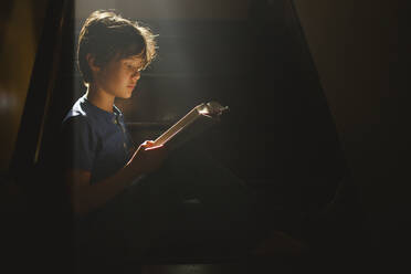 A boy sits on a darkened staircase in a bright beam of light reading - CAVF79566