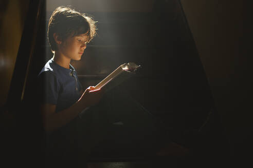 Ein Junge sitzt auf einer abgedunkelten Treppe in einem hellen Lichtstrahl und liest - CAVF79566