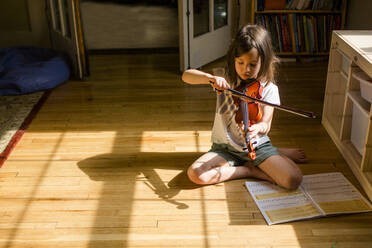 Ein kleines Mädchen sitzt auf dem Boden und spielt Geige vor einem aufgeschlagenen Notenbuch - CAVF79568