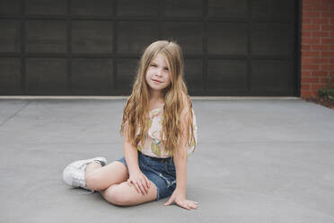 Porträt eines süßen jungen Mädchens, das vor einem Garagentor sitzt - CAVF79703