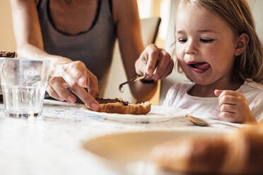 Porträt eines kleinen Mädchens, das seine Mutter dabei beobachtet, wie sie Schokoladencreme auf ein Croissant streicht - WFF00374