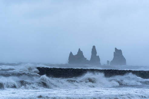 Island, Hofn, Meereswellen, die gegen eine Steinmauer an der Küste plätschern, mit Stapelfelsen im Hintergrund - DAMF00407