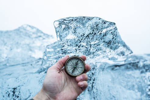 Iceland, Hand of man holding navigational compass in front of large block of ice - DAMF00408