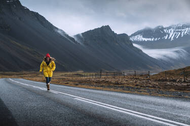 Island, Frau in gelbem Mantel läuft auf abgelegener isländischer Landstraße - DAMF00414