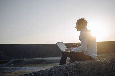 Älterer Geschäftsmann mit Laptop auf einer stillgelegten Minenhalde bei Sonnenuntergang - JOSEF00363