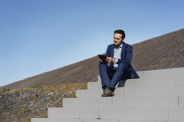 Älterer Geschäftsmann sitzt auf einer Treppe auf einer stillgelegten Bergwerkshalde und benutzt ein Tablet - JOSEF00374