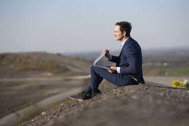 Mature businessman with laptop sitting on a disused mine tip - JOSEF00430