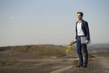 Mature businessman holding bunch of flowers and laptop on a disused mine tip - JOSEF00432