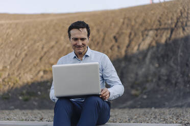 Älterer Geschäftsmann mit Laptop auf einer stillgelegten Bergwerkshalde - JOSEF00450