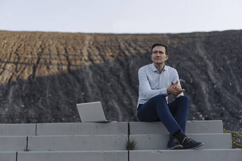 Älterer Geschäftsmann sitzt auf einer Treppe auf einer stillgelegten Minenhalde neben einem Laptop, lizenzfreies Stockfoto