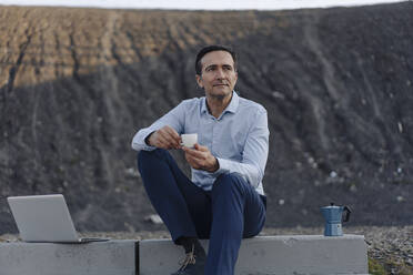 Mature businessman sitting on stairs on a disused mine tip next to laptop having a coffee break - JOSEF00453