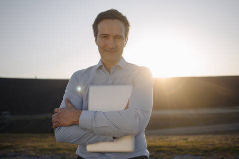 Porträt eines reifen Geschäftsmannes mit Laptop auf einer stillgelegten Minenhalde bei Sonnenuntergang, lizenzfreies Stockfoto