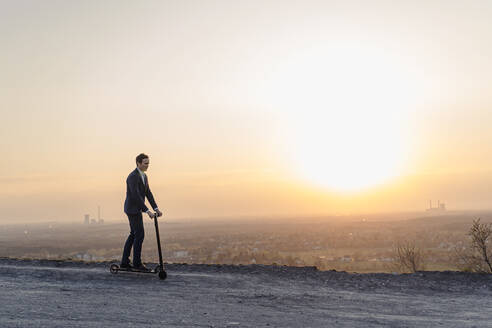 Älterer Geschäftsmann fährt auf einem Kick-Scooter auf einer stillgelegten Minenhalde bei Sonnenuntergang - JOSEF00489