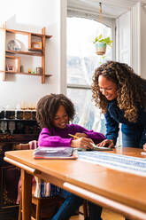 Smiling mother assisting daughter in doing homework at table in living room - CAVF79902