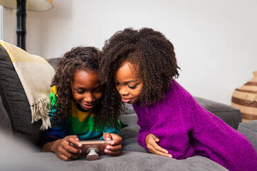 Mädchen schaut auf ihre Schwester, die auf dem Sofa zu Hause ein Handy benutzt - CAVF79908