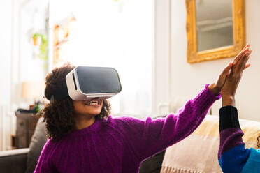 Happy girl high-fiving with sister while wearing virtual reality simulator at home - CAVF79909