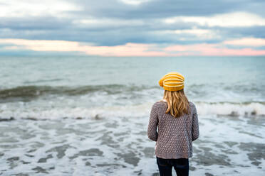 Tween girl wearing yellow knit hat looking at ocean at dusk - CAVF80006