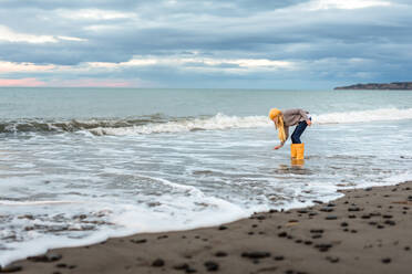 Jugendlicher mit gelben Stiefeln, der eine Muschel vom neuseeländischen Strand aufhebt - CAVF80007