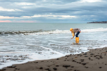 Preteen with yellow boots picking up shell from New Zealand beach - CAVF80007