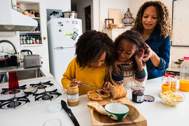Mutter bindet die Haare ihrer Tochter und schaut auf ihre Schwester, die in der Küche das Frühstück vorbereitet - CAVF80021