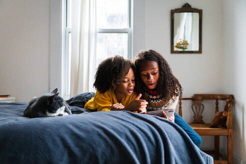 Mädchen zeigt ihrer Schwester das Smartphone, während sie zu Hause neben der Katze auf dem Bett liegt - CAVF80023
