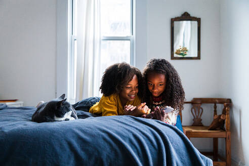 Lächelnde Mädchen, die ein Mobiltelefon benutzen, während sie neben einer Katze auf dem Bett liegen - CAVF80026