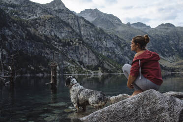 Frau mit Hund, die während eines Ausflugs auf einem Felsen an einem See sitzt. - CAVF80070