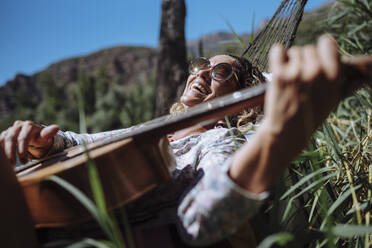 Frau mit Sonnenbrille lachend und Gitarre spielend auf einer Hängematte liegend - CAVF80080