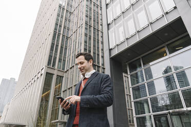 Lächelnder Geschäftsmann, der eine Textnachricht auf seinem Smartphone sendet, während er an einem Gebäude in Frankfurt, Deutschland, steht - AHSF02340