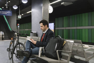 Geschäftsmann mit Fahrrad, der auf einer Bank am Bahnsteig einer U-Bahn-Station sitzt und Dokumente liest, Frankfurt, Deutschland - AHSF02345