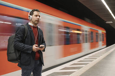 Businessman listening music through mobile phone while standing on platform against subway train - AHSF02348