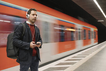 Geschäftsmann hört Musik über sein Handy, während er auf dem Bahnsteig vor einem U-Bahn-Zug steht - AHSF02348