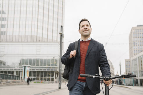 Lächelnder Geschäftsmann, der wegschaut, während er mit dem Fahrrad auf einer Straße in Frankfurt, Deutschland, spazieren geht - AHSF02354