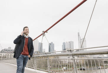 Smiling businessman talking on mobile phone while walking on bridge against sky in Frankfurt, Germany - AHSF02359