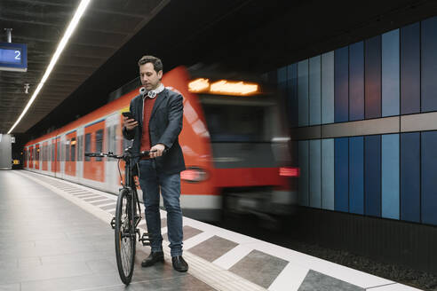 Businessman with bicycle text messaging on smart phone while standing on platform against subway train - AHSF02364