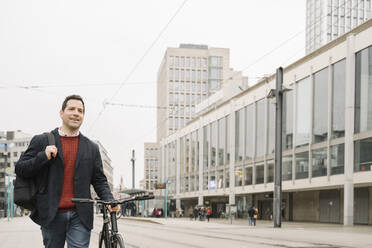 Lächelnder Unternehmer, der wegschaut, während er mit dem Fahrrad auf der Straße in Frankfurt, Deutschland, spazieren geht - AHSF02367