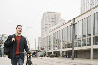 Smiling entrepreneur looking away while walking with bicycle on road in Frankfurt, Germany - AHSF02367